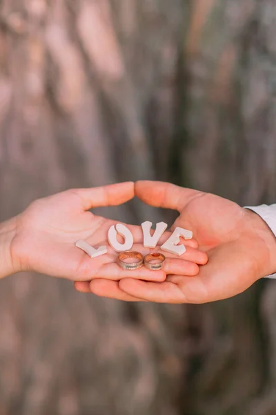 Anillos de boda dorados, letras de madera en las palmas de los novios, primer plano — Foto de Stock