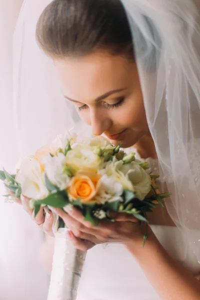 Прекрасная молодая невеста, стоящая в белой комнате и нюхающая свадебный букет. — стоковое фото