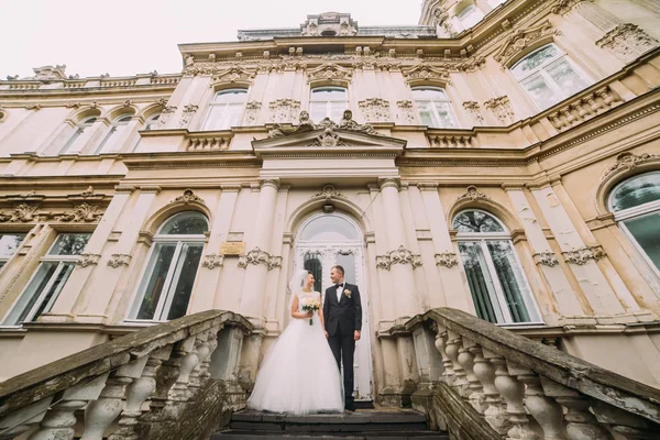 Bela noiva em véu branco e belo noivo em pé nas escadas contra o pano de fundo do edifício antigo — Fotografia de Stock