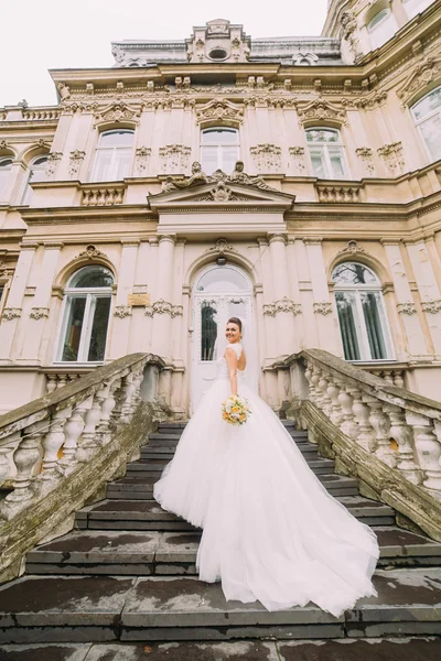 Beautiful bride in wedding dress with long train standing back on the stairs — Stock Photo, Image