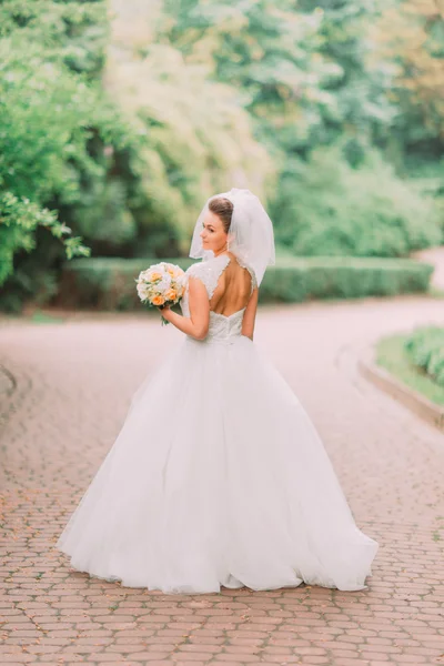 Красива чуттєва молода брюнетка наречена в білій весільній сукні і вуаль, що тримає букет на відкритому повітрі на природному фоні . — стокове фото