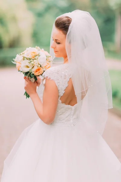 Linda sensual jovem noiva morena em vestido de noiva branco e véu segurando buquê ao ar livre no fundo natural, close-up — Fotografia de Stock