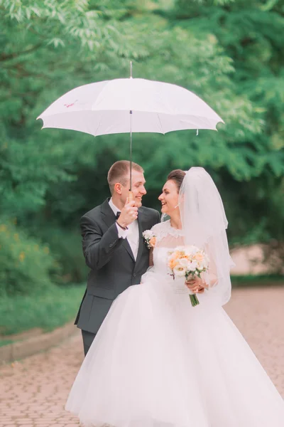 Happy wedding couple walking outdoors in park with white umbrella in rainy weather. — Stock Photo, Image