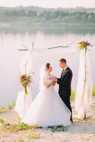 Hermosa novia en vestido blanco y novio guapo con traje negro de pie cogido de la mano bajo arco en la ceremonia de boda de playa cerca del lago —  Fotos de Stock