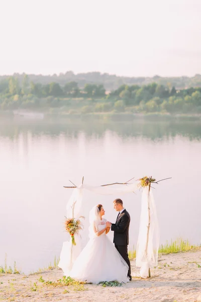 Hermosa novia en vestido blanco y novio guapo con traje negro de pie cogido de la mano bajo arco en la ceremonia de boda de playa cerca del lago —  Fotos de Stock