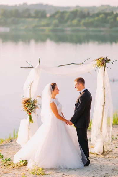 Hermosa novia en vestido blanco y novio guapo con traje negro de pie cogido de la mano bajo arco en la ceremonia de boda de playa cerca del lago —  Fotos de Stock