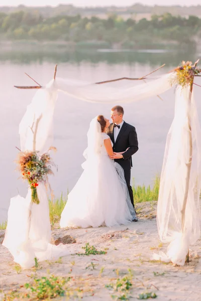 Hermosa novia en vestido blanco y novio guapo con traje negro abrazando bajo arco en la ceremonia de boda de playa cerca del lago —  Fotos de Stock