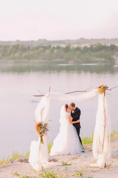 Красива наречена в білій сукні і красивий наречений в чорному костюмі цілується під аркою на пляжній весільній церемонії біля озера — стокове фото
