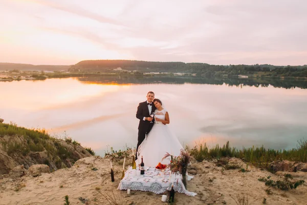 Novia en velo blanco y novio con traje negro abrazándose cerca de la mesa romántica en la playa al atardecer — Foto de Stock