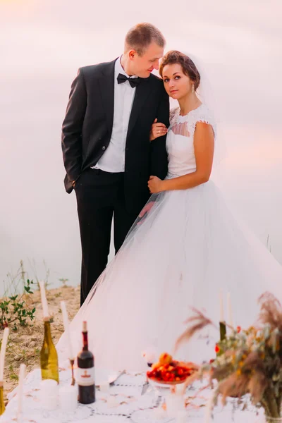 Novia en velo blanco y novio con traje negro abrazándose cerca de la mesa romántica en la playa al atardecer —  Fotos de Stock