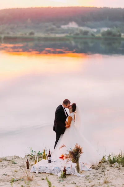 Sposa in velo bianco e sposo indossa abito nero baciarsi vicino al romantico tavolo da pranzo sulla spiaggia al tramonto — Foto Stock