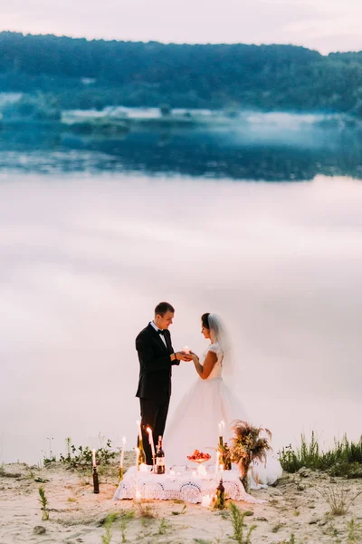 Braut in weißem Schleier und Bräutigam im schwarzen Anzug küssen sich am romantischen Esstisch am Strand bei Sonnenuntergang — Stockfoto