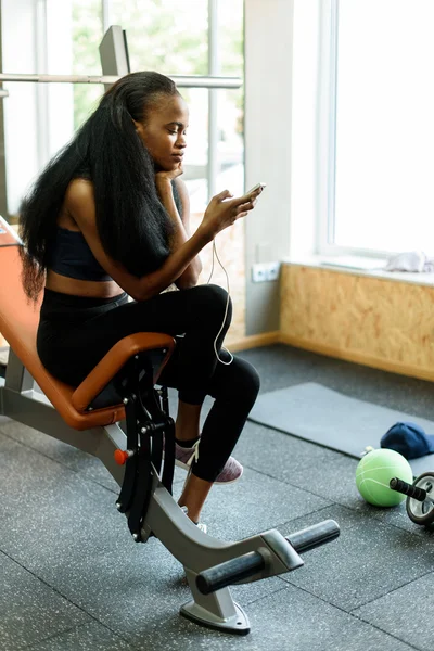Портрет очаровательной черной молодой женщины с роскошными длинными волосами, переписывающейся на смартфоне в спортзале — стоковое фото