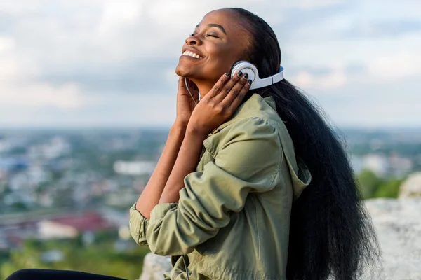 Close-up portrait of happy smiling young black african american woman listening to music. Blurred cityscape on background — Stock Photo, Image