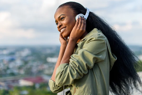Close-up portrait of gorgeous young black african american woman listening to music. Blurred cityscape on background — Stock Photo, Image