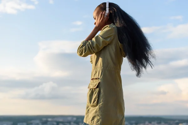 Belle jeune femme noire américaine souriante aux cheveux longs écoutant de la musique. Paysage urbain flou en arrière-plan — Photo
