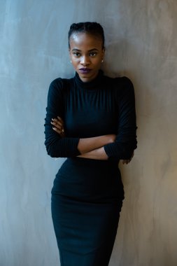 Kolları katlanmış gri arka plan üzerinde duran ve kameraya bakarak ciddi bir Afrikalı ya da siyah amerikalı kadın portresi