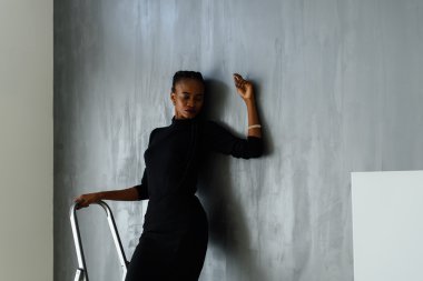 Güzel ve seksi siyah kadın stüdyoda gri duvara yaslanmış el aşağı seyir poz koyu elbiseli