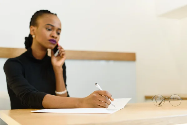 Serious confident young African or black American business woman on phone taking notes in office — Stock Photo, Image