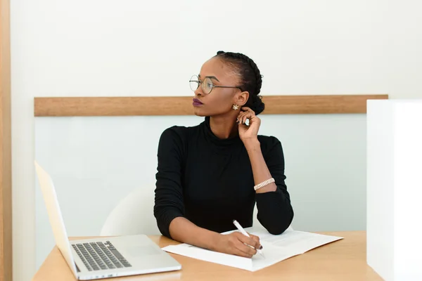 Pensativa mujer de negocios negra en gafas tocándose el cuello mirando hacia el escritorio con bloc de notas y portátil en la oficina —  Fotos de Stock