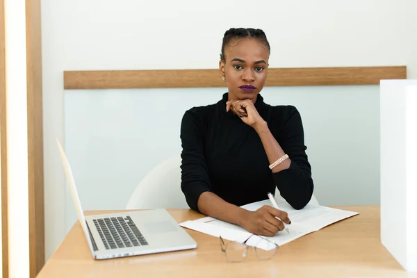 Elegante mujer africana o negra americana sosteniendo la barbilla trabajando en el escritorio con bloc de notas y computadora portátil en la oficina —  Fotos de Stock