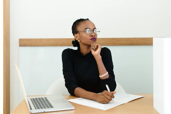 Confident black business woman in glasses holding chin looking away at desk with notepad and laptop in office — Stock Photo, Image