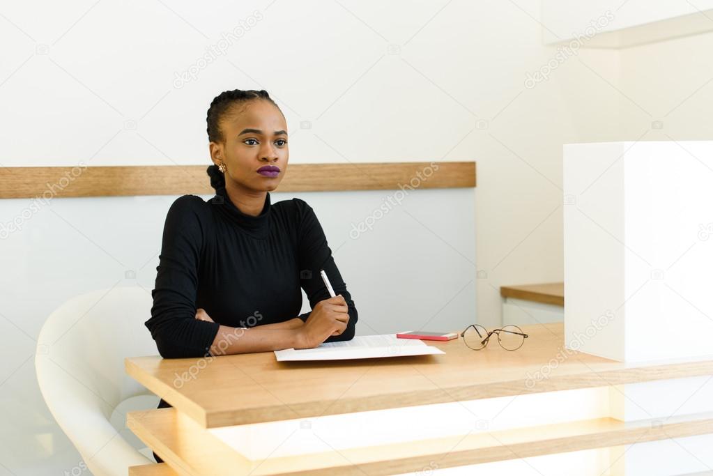 Confident young black business woman holding pen and thinking at desk in office