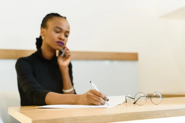 Sério confiante jovem Africano ou negro mulher de negócios americano no telefone tomar notas no escritório — Fotografia de Stock