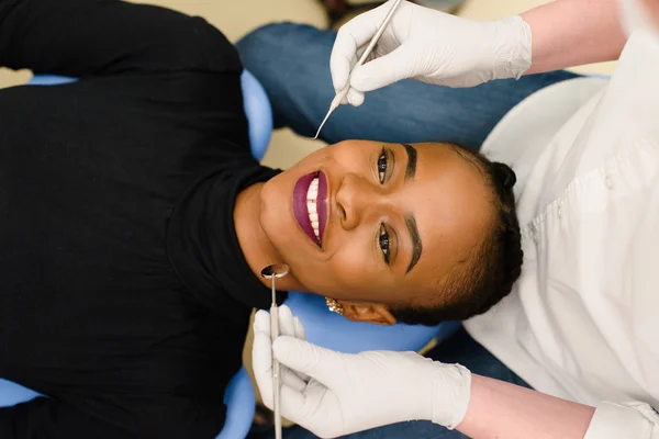 Молодая афро-американская этническая черная женщина улыбается, а стоматолог в белых латексных перчатках проверяет состояние зубов — стоковое фото