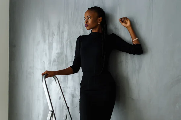 Hermosa y sexy mujer negra en vestido oscuro posando mirando hacia otro lado apoyando la mano en la pared gris en el estudio — Foto de Stock