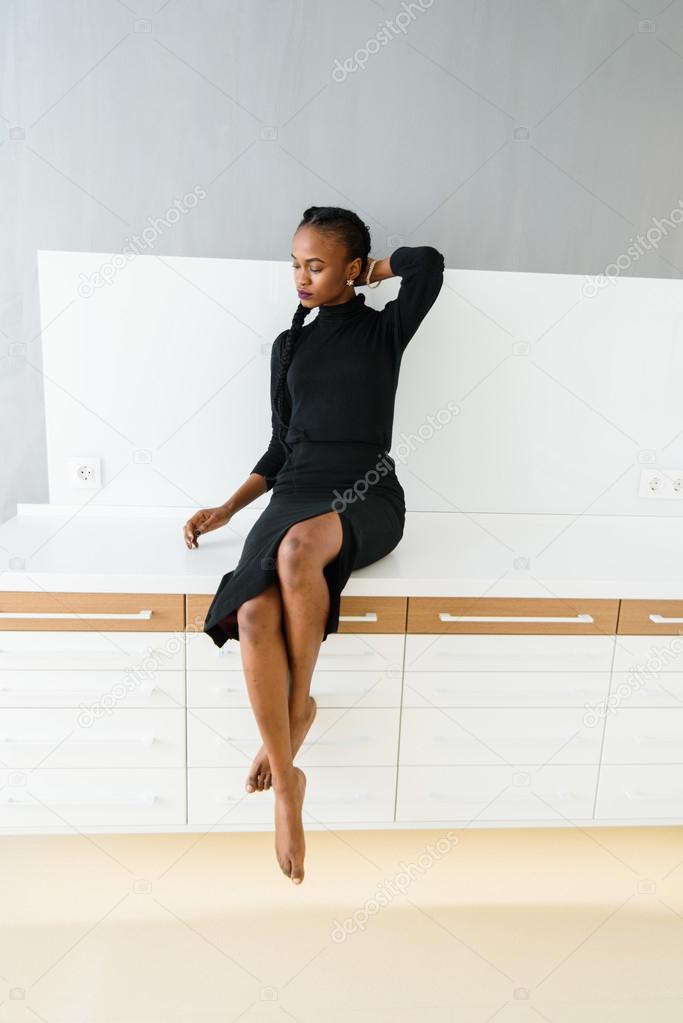 Elegant african or black american woman in dark dress sitting on desk in light interior
