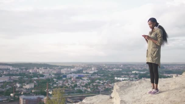 Lovely young black lady in cap choose music to listen in her smartphone. Blurred cityscape background — Stock Video