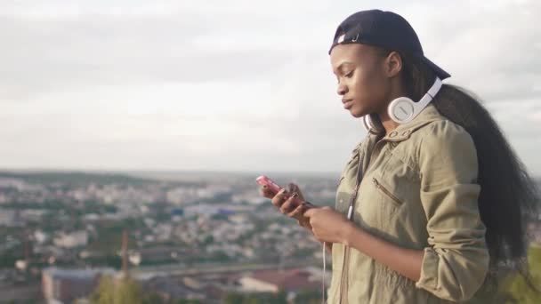 Menina afro-americana bonito em cap escolher a música para ouvir em seu smartphone. Cenário urbano desfocado — Vídeo de Stock