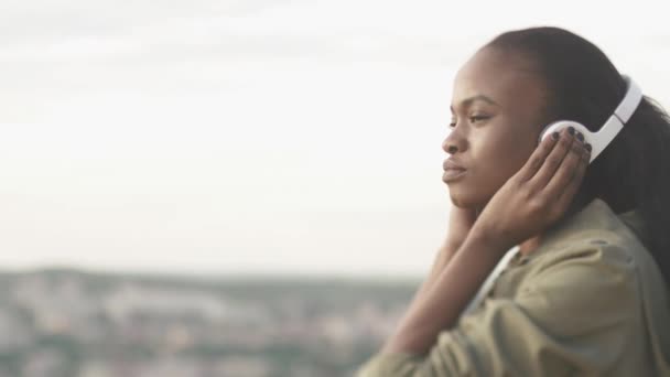 Retrato de cerca de una hermosa dama negra escuchando música en auriculares sobre un fondo borroso de la ciudad. Momento de verdadero relax — Vídeo de stock