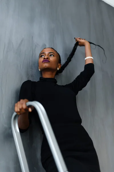 Young pretty black american woman touching her thick plait looking up on dark studio background