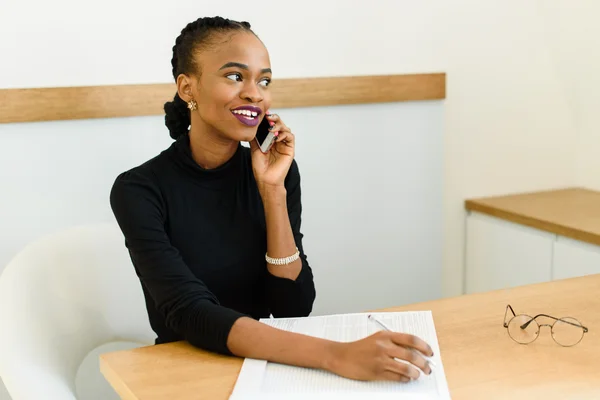Sorrindo jovem mulher de negócios preto no telefone tomar notas um olhar para cima no escritório — Fotografia de Stock