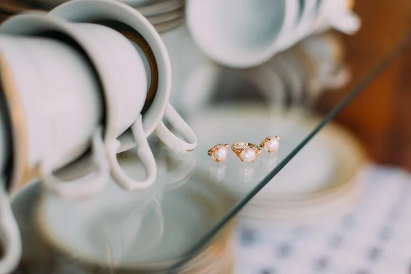 Extreme close-up golden finger ring and earrings embellished with pearls on glass cupboard shelf. Porcellain cups, plates at background