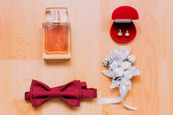 Conjunto de acessórios masculinos e femininos sobre fundo de madeira rústica. Perfume, borgonha laço, caixa vermelha com pérola decorados brincos boutonniere floral branco — Fotografia de Stock
