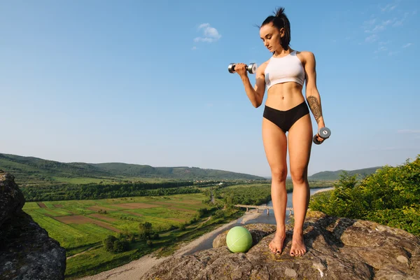 Sexy brunette vrouw fitness instructeur doen oefeningen met halters en bal. Buiten trainingsconcept. Groene bos bergen achtergrond — Stockfoto