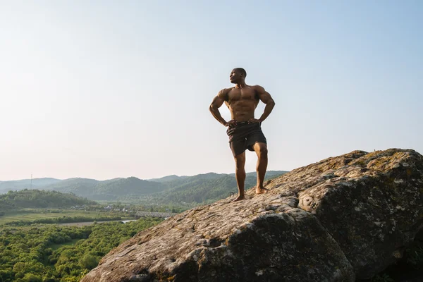 Portret van sterke zwarte Afro-Amerikaanse man bodybuilder met naakte torso poseren op de rotsachtige piek. Blauwe bewolkte hemelachtergrond — Stockfoto