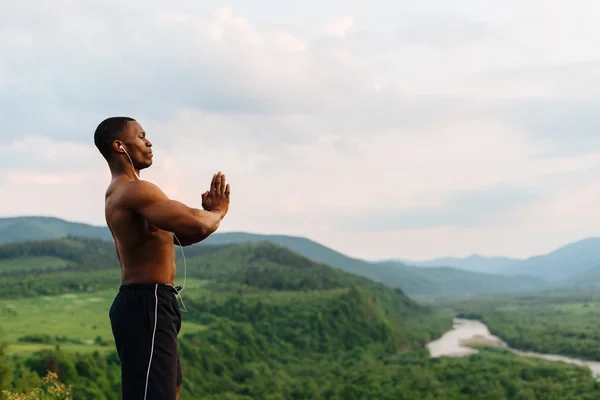 Silhouette eines schwarzafrikanisch-amerikanischen muskulösen Mannes mit erhobenen Händen vor wunderschönem Berghintergrund. Amen pray Konzept. — Stockfoto