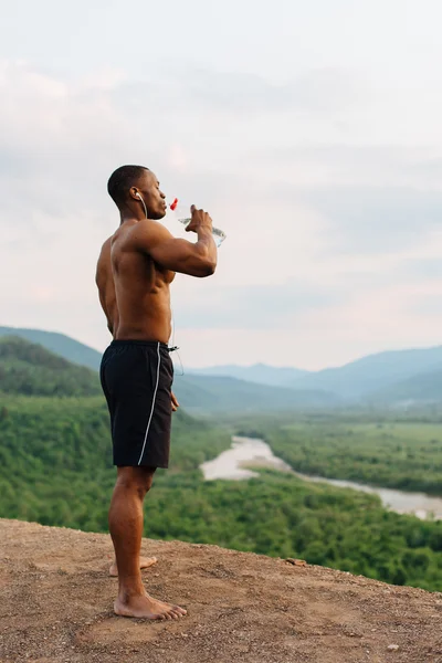 Full-length Portret van Afrikaanse Amerikaanse gespierde man drinkwater na sporttrainingen. Adembenemende groene berglandschap op achtergrond — Stockfoto