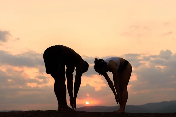 Silhuetas de casal raça mista ajuste alongamento e treinamento juntos no fundo das montanhas rochosas com pôr do sol dourado. Conceito de aptidão — Fotografia de Stock