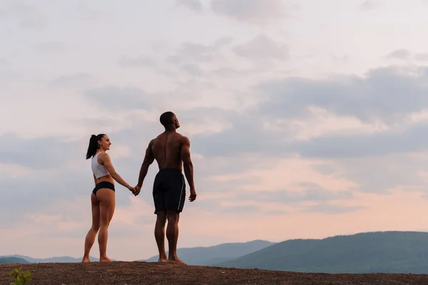 Momento de armonía. Feliz pareja de raza mixta atlética mujer caucásica con cuerpo afroamericano tomados de la mano y viendo el amanecer juntos en el pico de la montaña — Foto de Stock