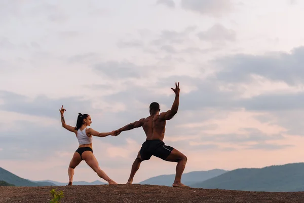 Gemengde ras gymnastische paar met perfecte lichamen in sportkleding dansen op bergen landschap achtergrond. Roze avondrood en witte wolken — Stockfoto