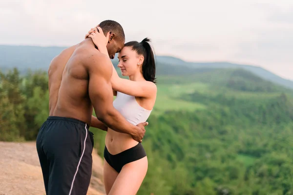 Porträtt av sexiga passa blandras par med perfekta kroppar i sportkläder mjukt omfamnar på berg landskap bakgrund. — Stockfoto