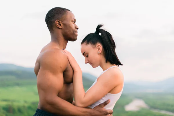 Vista laterale di sexy vestibilità mista coppia gara con corpi perfetti in abbigliamento sportivo dolcemente abbracciando su montagne paesaggio sfondo . — Foto Stock