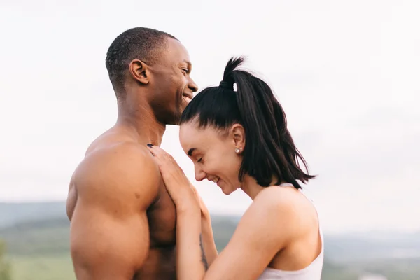 Närbild av sexiga passa blandras par med perfekta kroppar i sportkläder mjukt omfamnar på berg landskap bakgrund. — Stockfoto