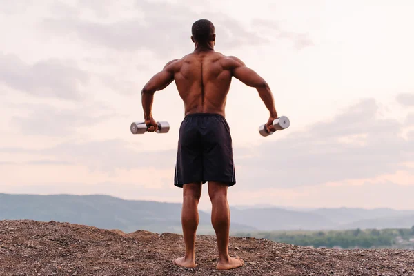 Полнометражный портрет африканского мускулистого атлета, поднимающего гантели на фоне закатного неба. Вид сзади — стоковое фото