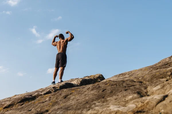Visão traseira do fisiculturista afro-americano bonito posando na rocha contra o céu azul nublado — Fotografia de Stock